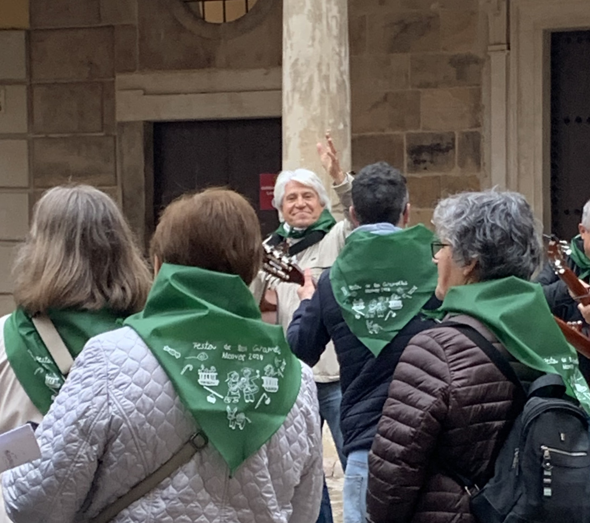 Entre las voces del coro con pañuelos verdes, Joan Marc sonríe con una mano levantada y la guitarra ante la fachada de piedra de un edificio.