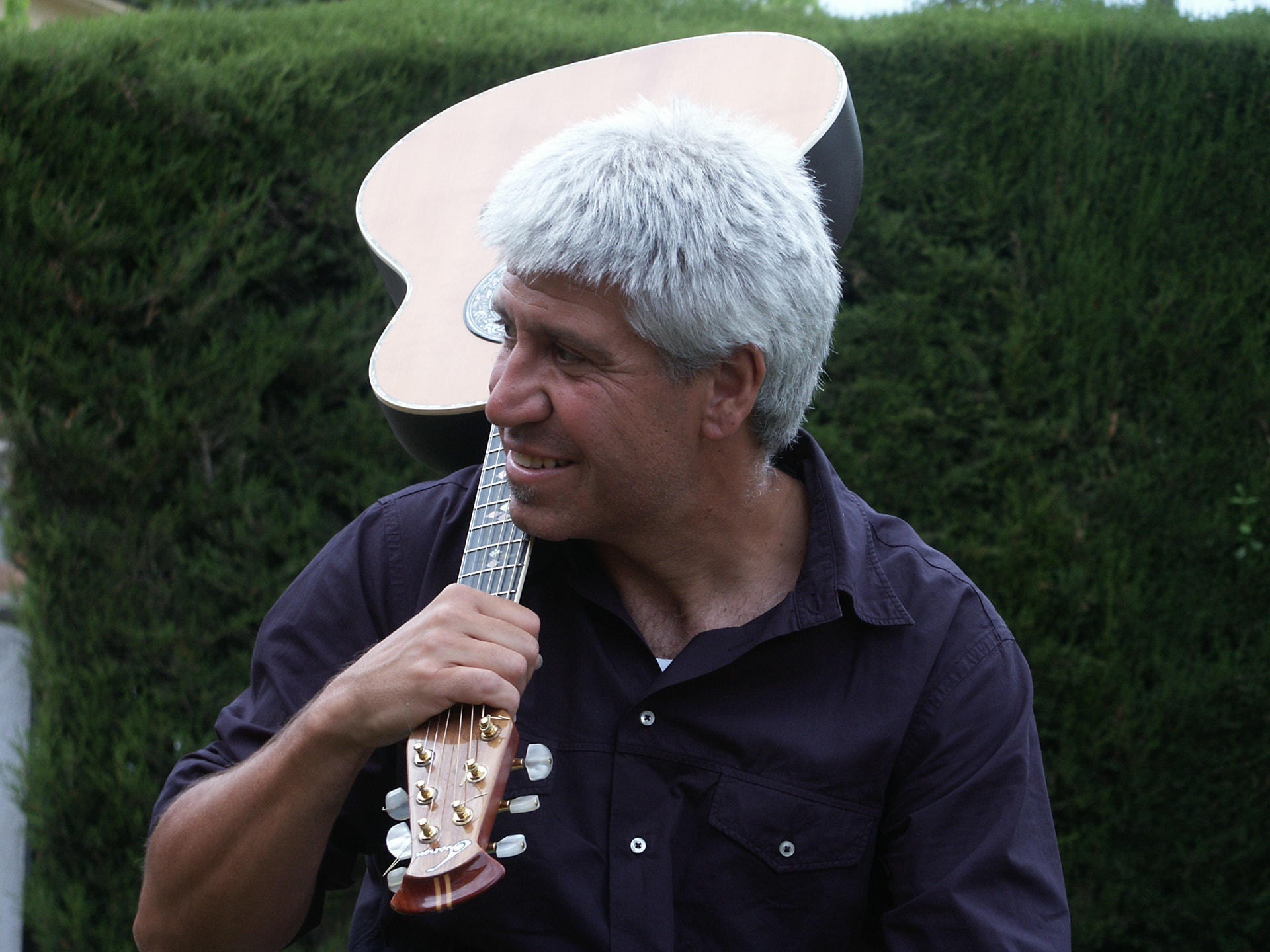 Retrato de Joan Marc con la guitarra al hombro, sonriendo ante un seto verde.