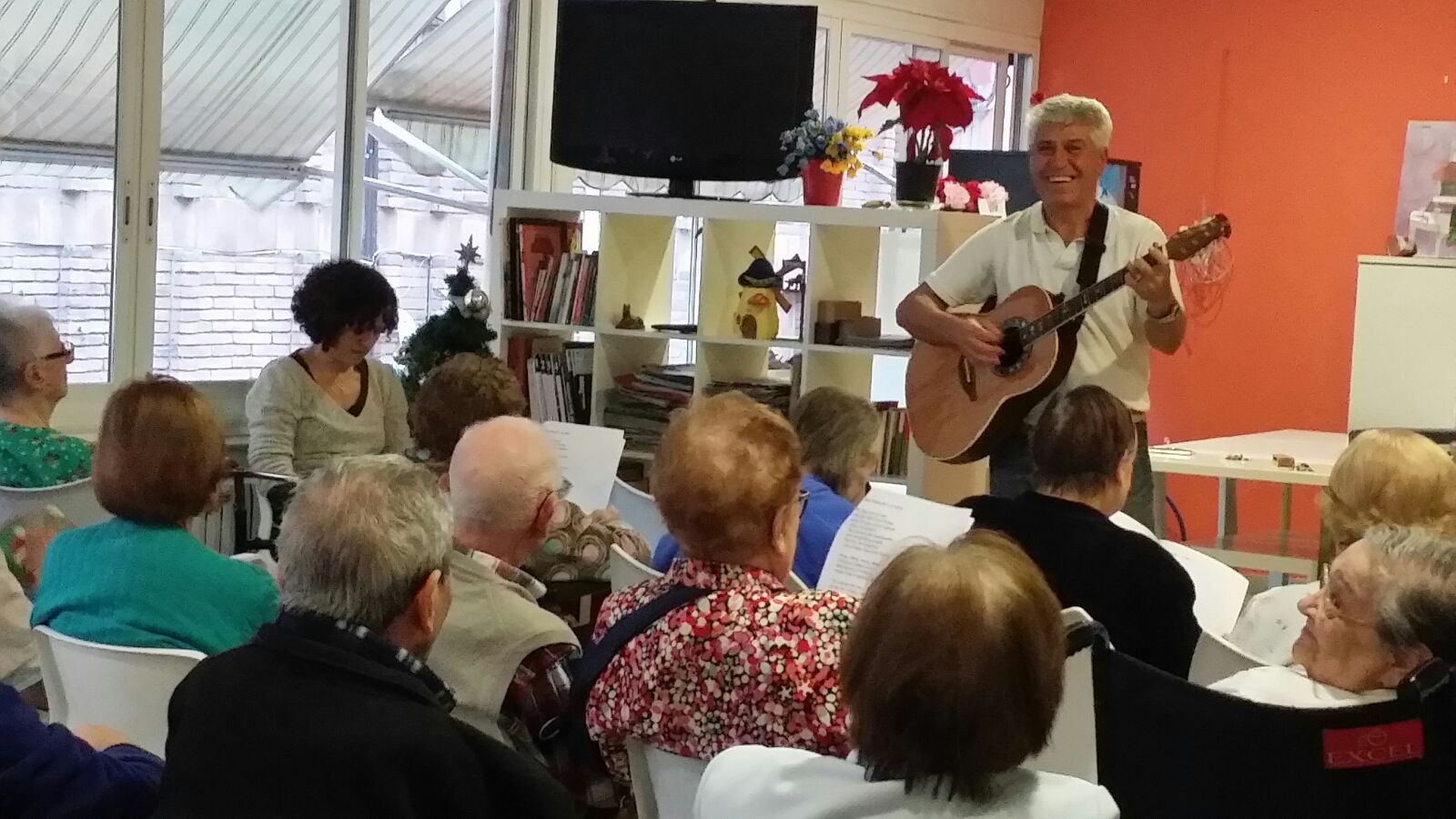 Joan Marc, de pie y sonriendo, con polo blanco y guitarra acústica, ante un grupo de personas mayores sentadas en una sala del centro.