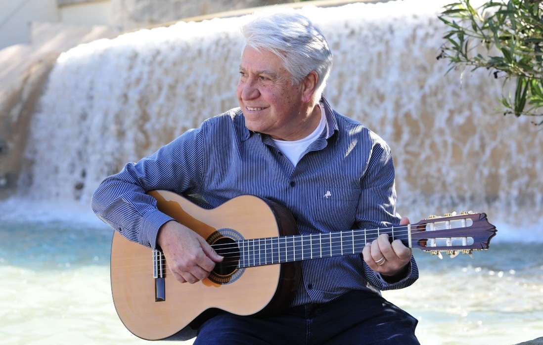 Joan Marc sonríe mirando hacia un lado mientras toca la guitarra clásica, sentado junto a una cascada de agua y rodeado de vegetación.