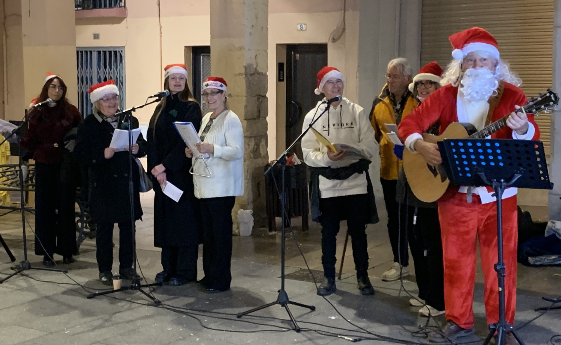 Nadales nocturnas bajo una columnata: cantores con gorros de Papá Noel y partituras y, a la derecha, Joan Marc tocando la guitarra vestido de Santa Claus.