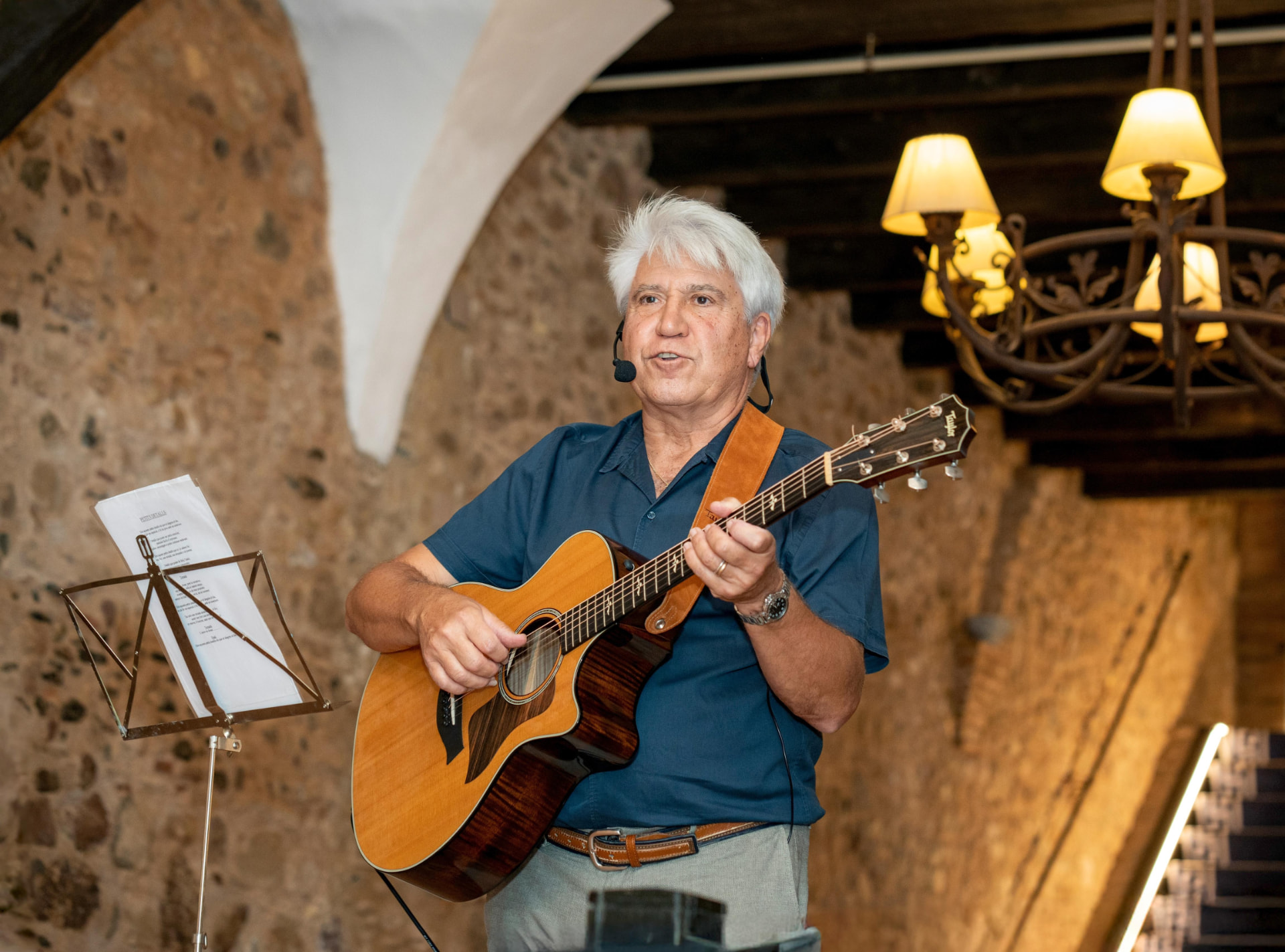 Joan Marc canta con micrófono de diadema y guitarra acústica en una sala de paredes de piedra, arcos blancos y lámparas de luz cálida.