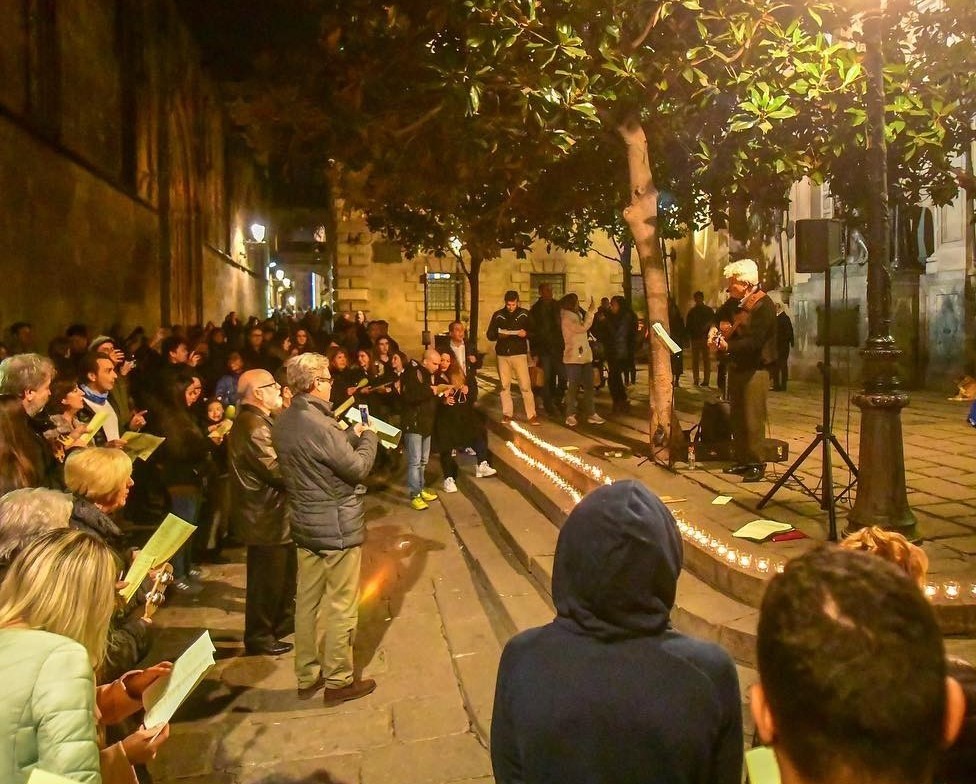 Joan Marc tocando y cantando ante una multitud que sigue las letras en una calle empedrada del Barri Gòtic, con velas encendidas.