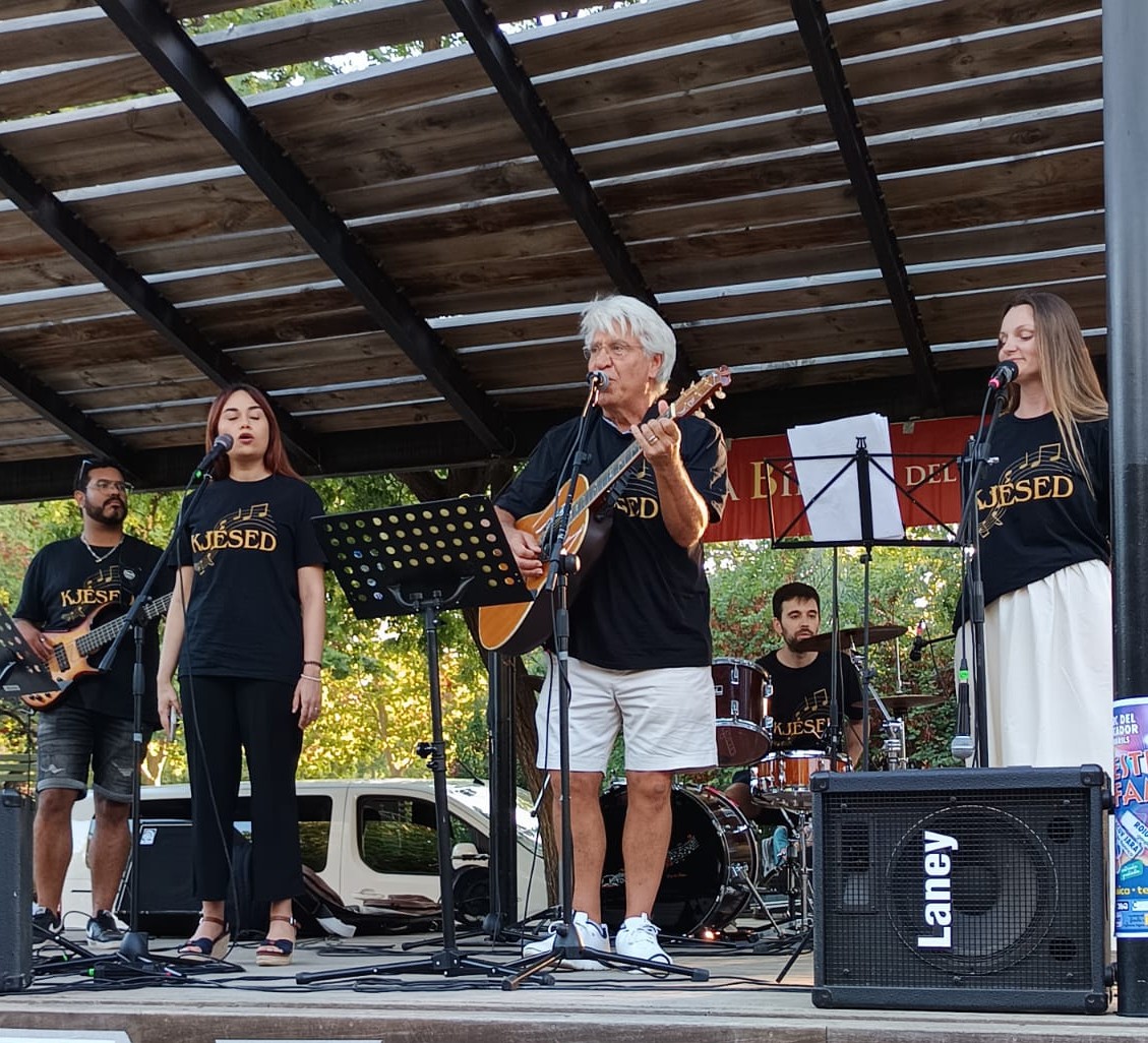 Joan Marc, con camiseta negra del grupo y pantalón blanco, toca la guitarra en el centro de la banda bajo un techo de madera, flanqueado por dos voces y la batería.