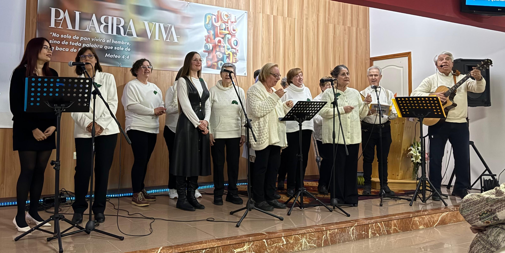 Un coro de una docena de voces, con blusas blancas y prendas negras, canta en un escenario de madera; a la derecha, Joan Marc toca la guitarra con micrófono de diadema.