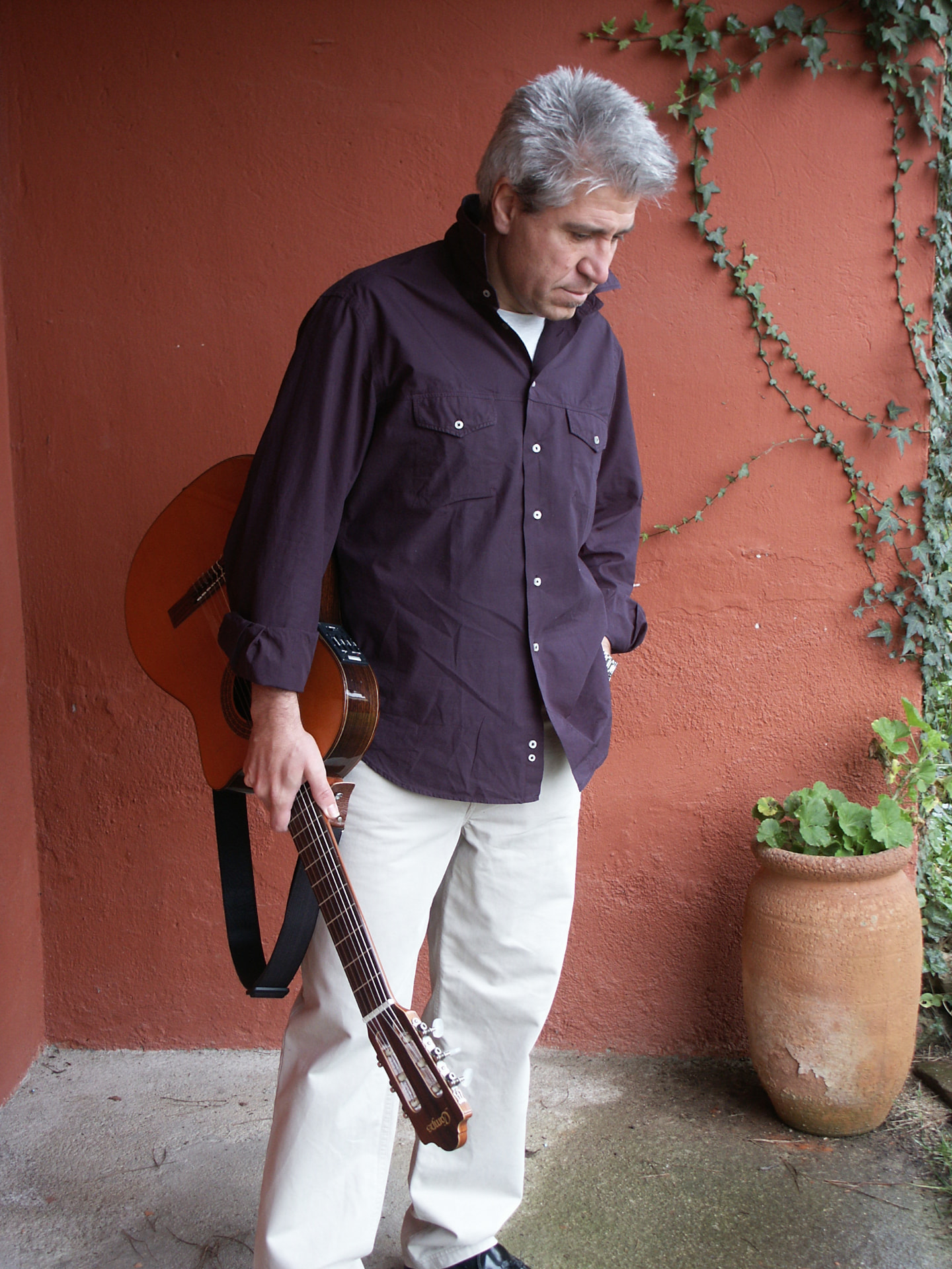 Joan Marc de pie con una guitarra en la mano, junto a una pared rojiza con hiedra y una maceta.
