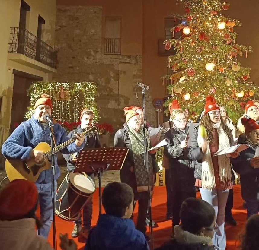 Joan Marc toca la guitarra junto a un grupo que canta con partituras y gorros navideños, ante un árbol de Navidad iluminado y el público de la plaza.