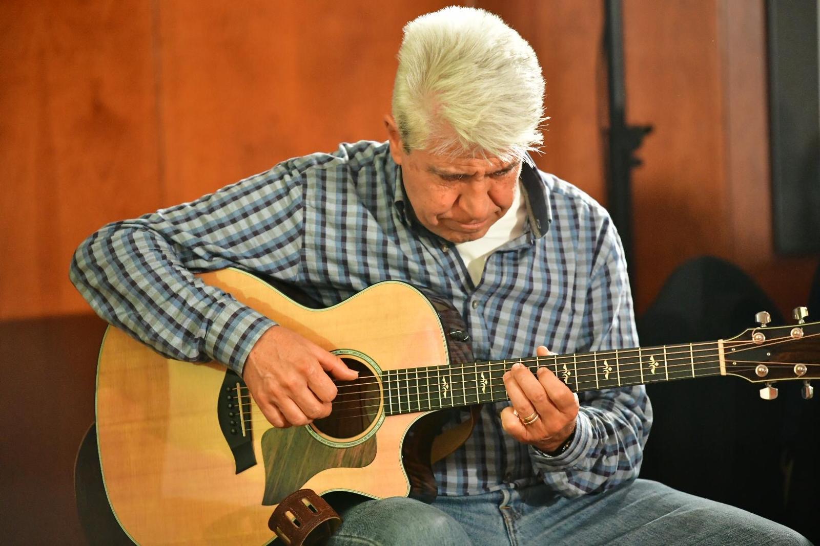 Primer plano de Joan Marc con camisa de cuadros, concentrado en la guitarra acústica sobre un fondo de madera cálida.