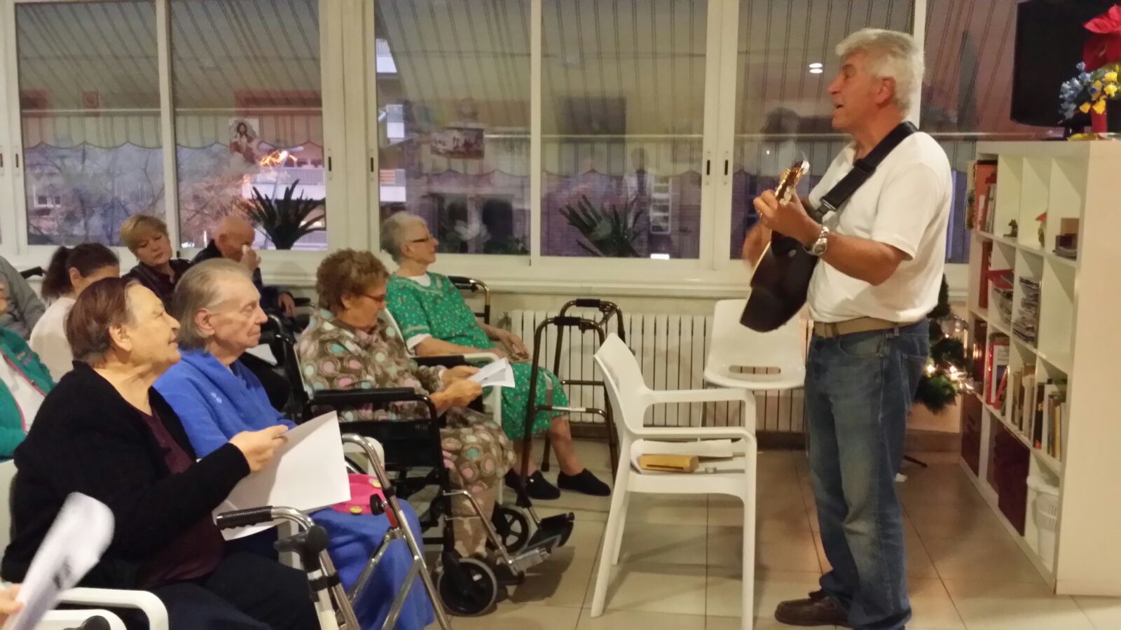 Joan Marc, de perfil, canta de pie ante mujeres mayores sentadas en sillas de ruedas, con hojas de canciones en las manos.