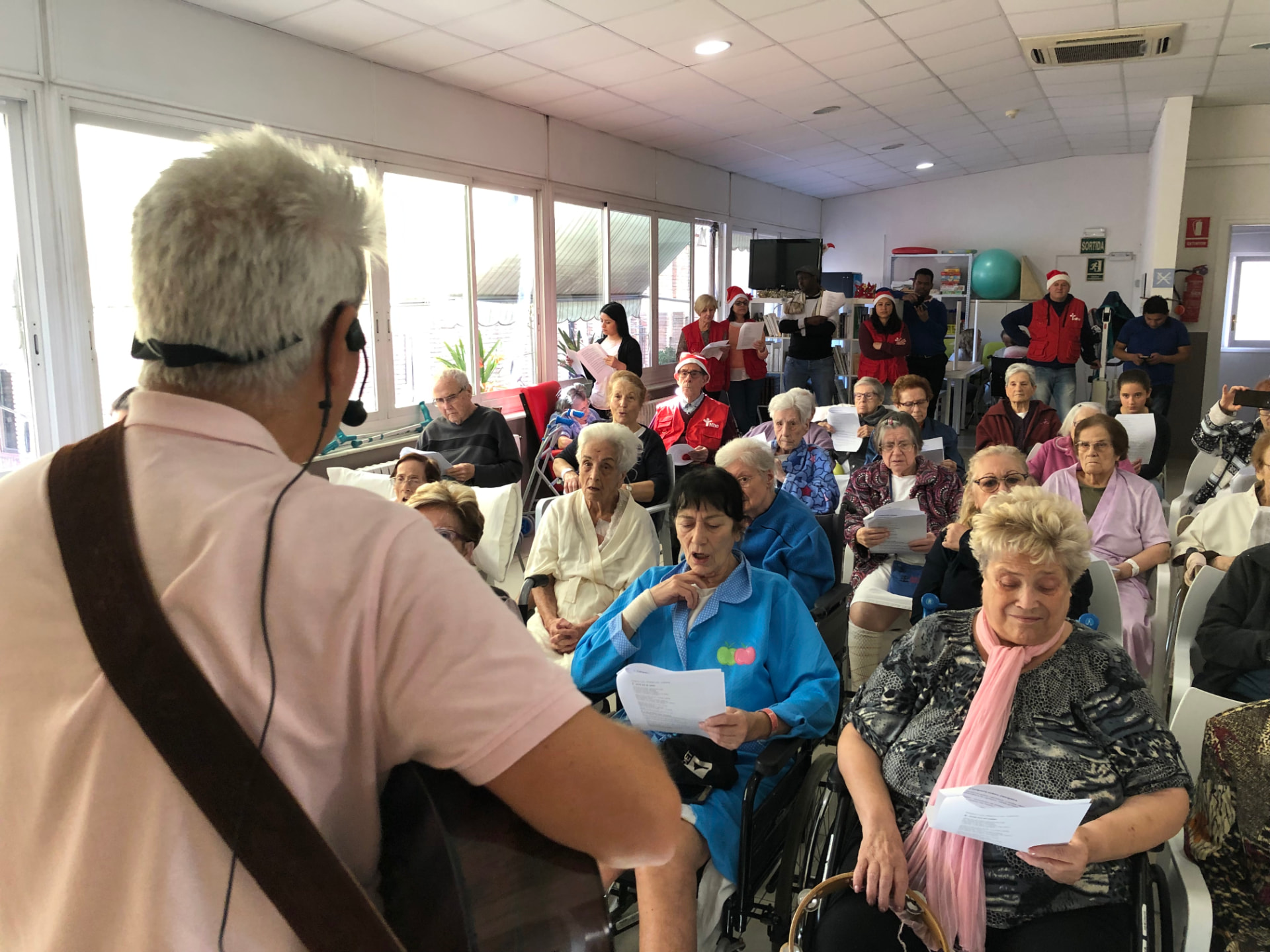 Joan Marc, visto desde atrás y con micrófono de diadema, canta ante el público del centro; al fondo, voluntarios con chalecos rojos y gorros de Papá Noel.