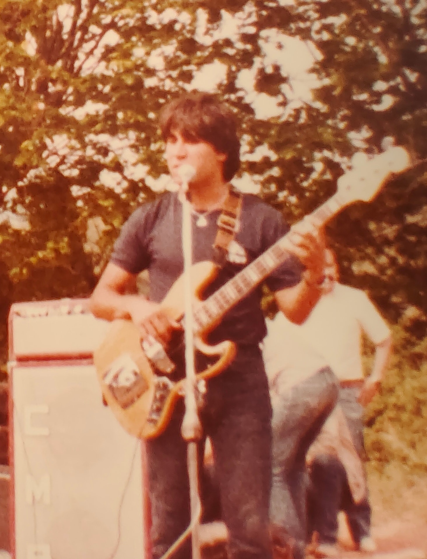 Joan Marc tocando un bajo eléctrico y cantando ante un micrófono, con árboles y amplificadores al fondo.
