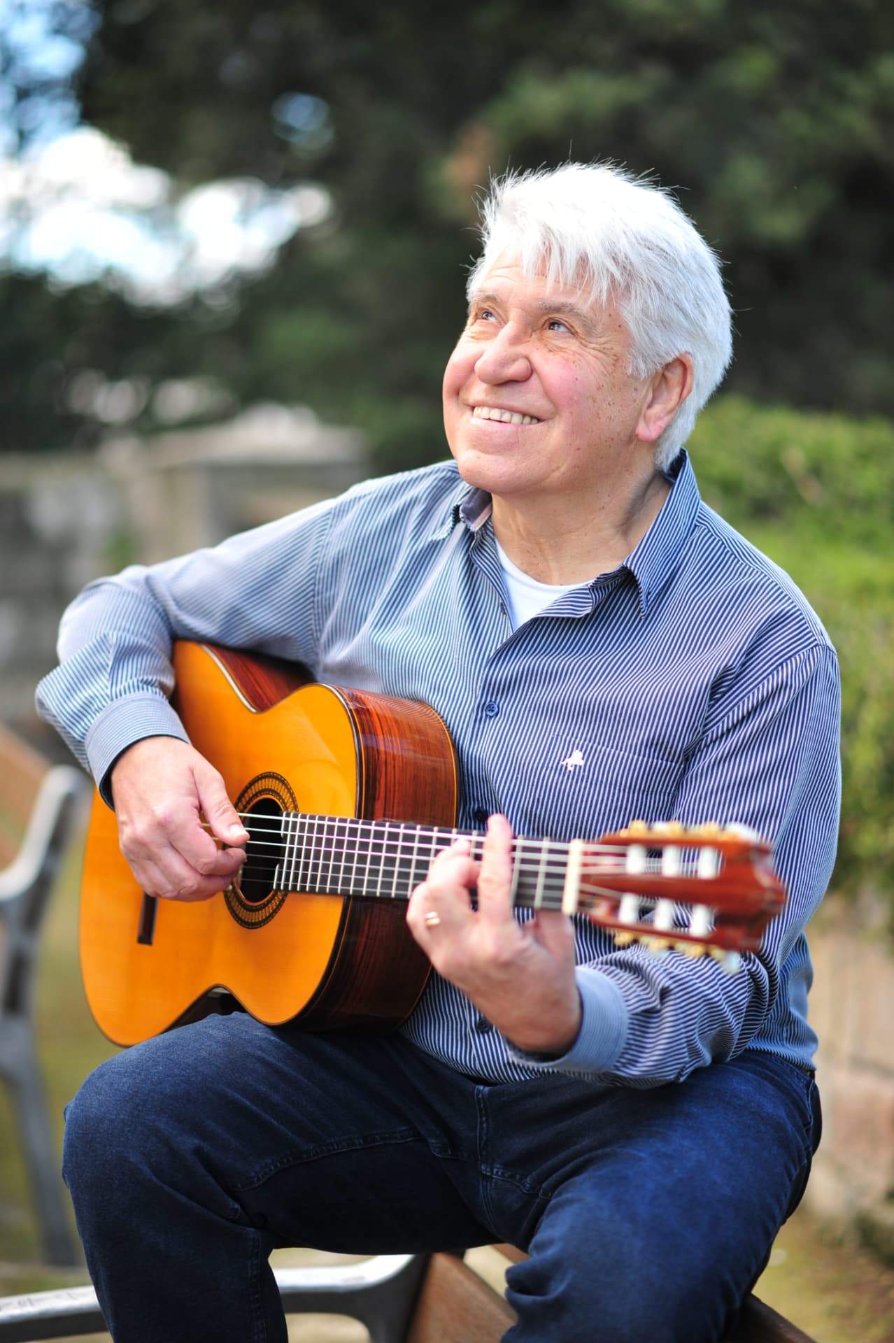 Sentado en un banco de un jardín, Joan Marc toca la guitarra clásica y mira hacia arriba sonriendo, con árboles al fondo.