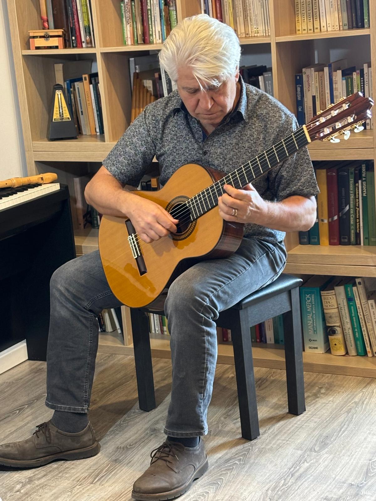 Joan Marc toca la guitarra clásica sentado ante una biblioteca de madera, junto a un piano, una flauta y un metrónomo.