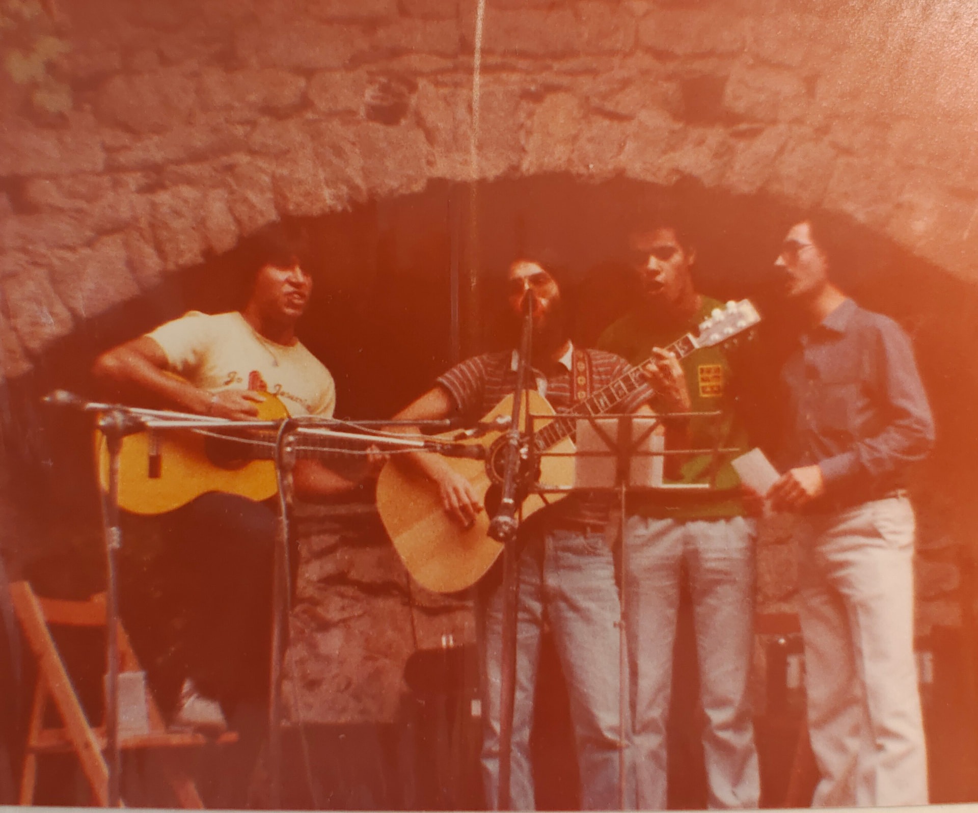 Cuatro hombres cantando y tocando la guitarra sobre un escenario con un arco de piedra al fondo.