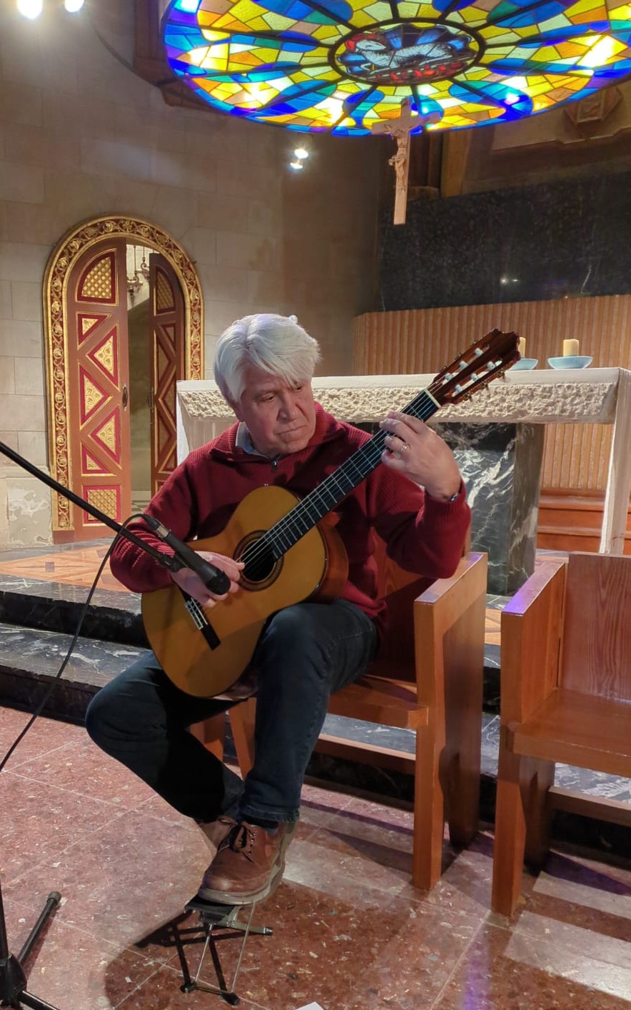 Joan Marc, sentado con jersey rojo, toca la guitarra clásica ante el altar de una capilla, bajo una vidriera circular de colores y un crucifijo.