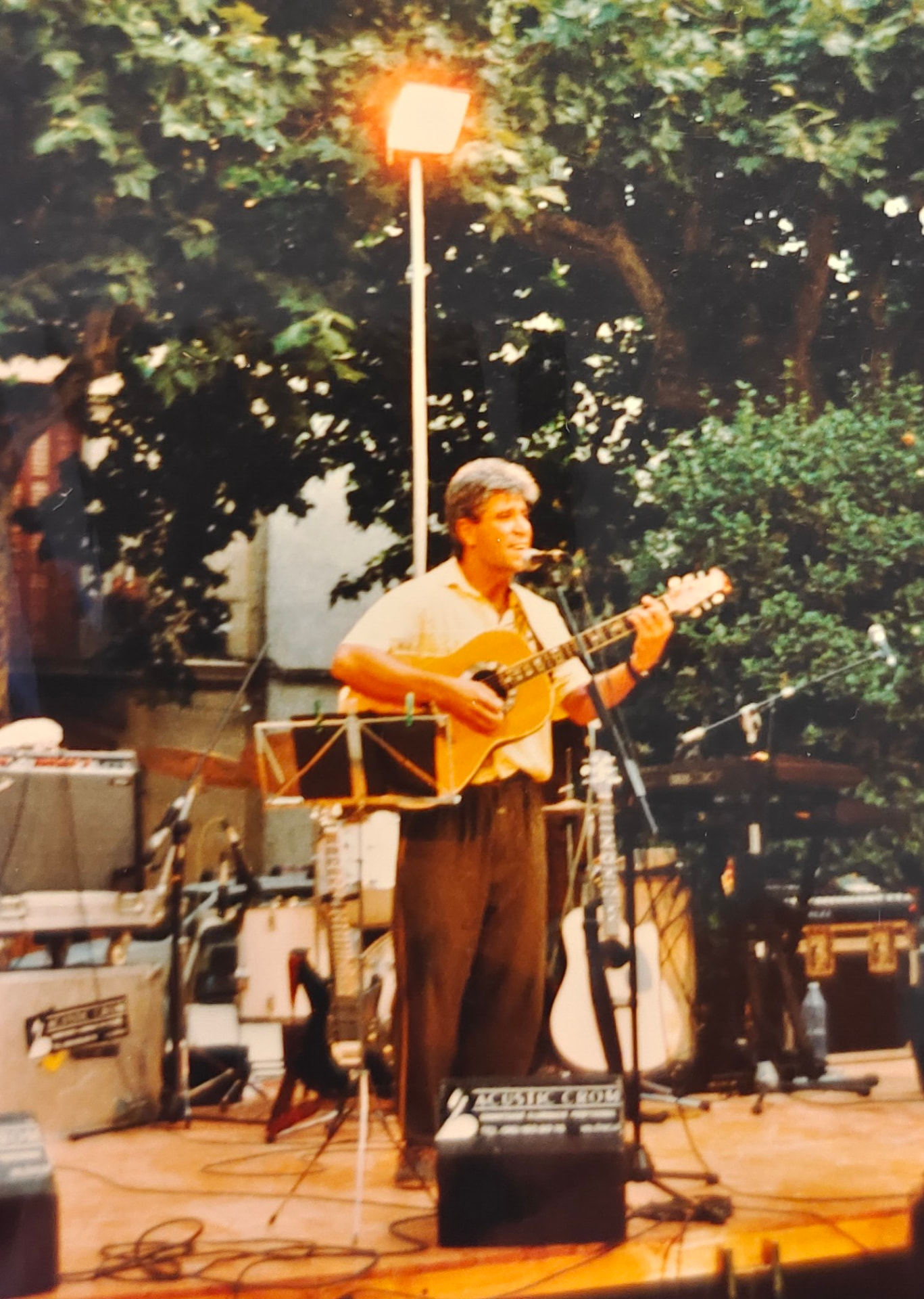Joan Marc cantando y tocando la guitarra acústica en un escenario al aire libre, con árboles y un atril detrás.