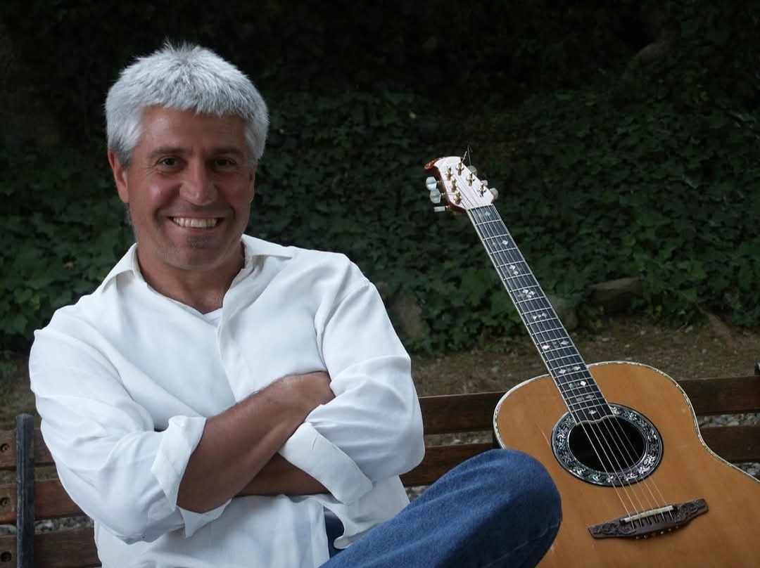 Joan Marc sentado en un banco de madera con los brazos cruzados y la guitarra a su lado, frente a una pared de hiedra.