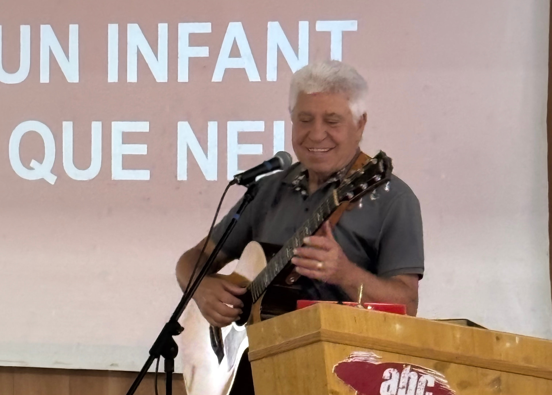 Primer plano de Joan Marc, con los ojos casi cerrados y una sonrisa, cantando y tocando la guitarra tras un atril de madera.