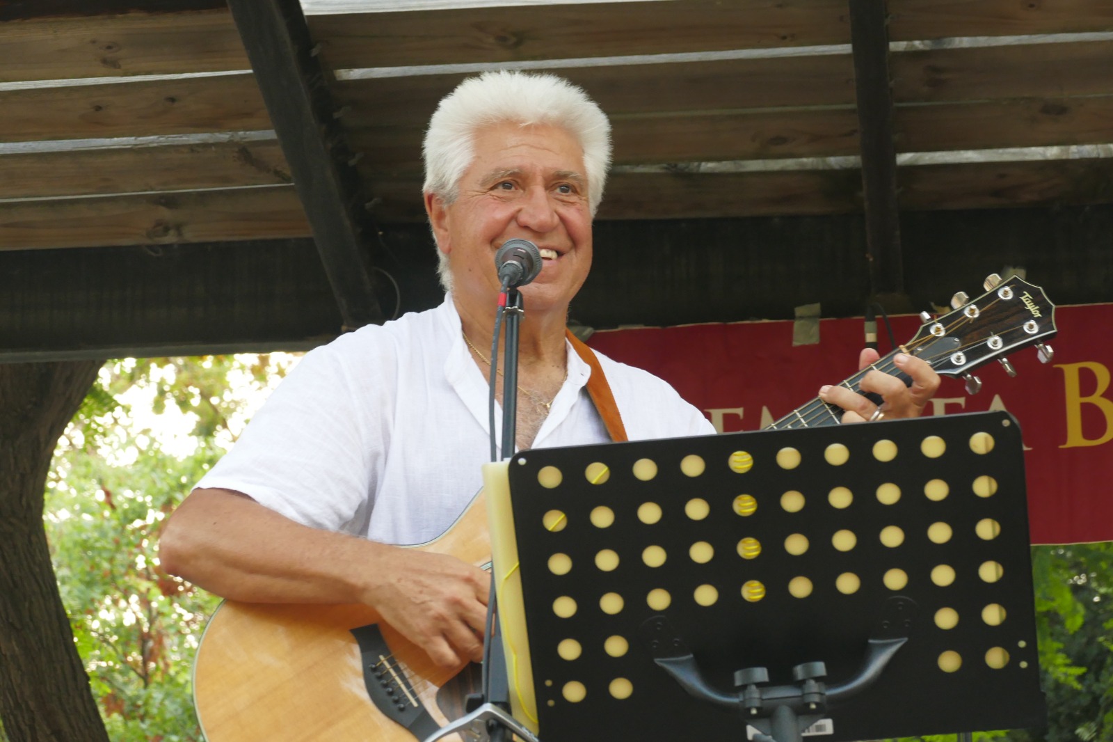 Joan Marc, con camisa blanca de lino, canta sonriendo al micrófono con la guitarra acústica bajo la pérgola de madera del escenario.