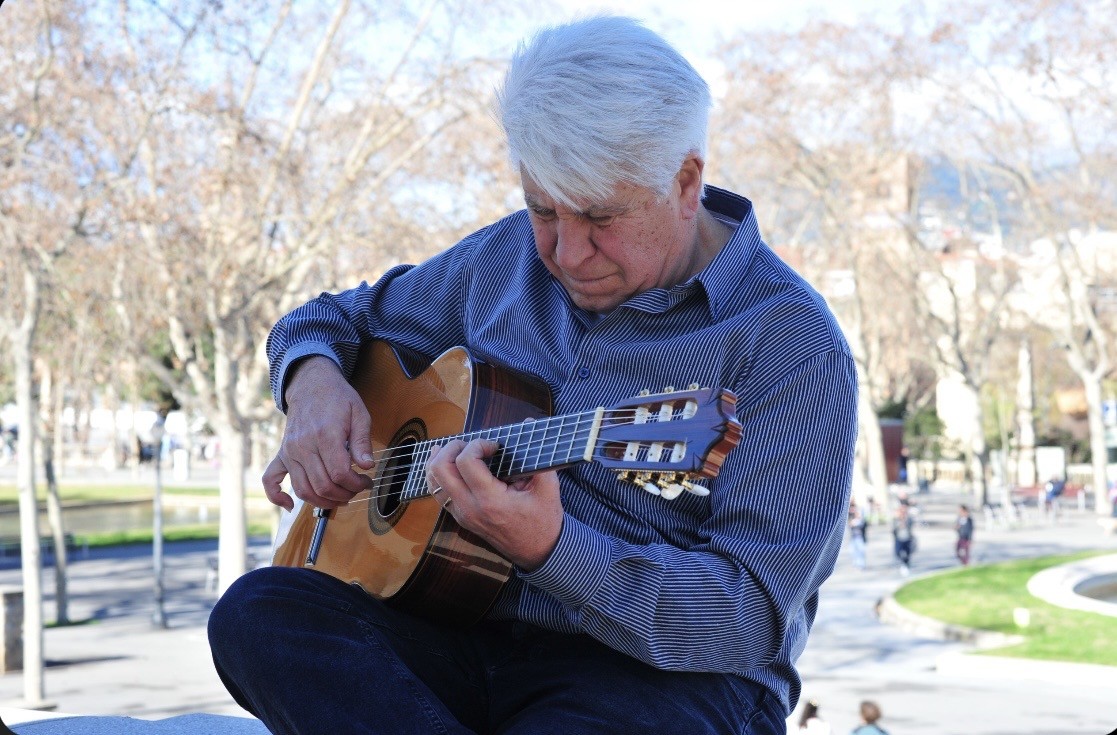 Joan Marc, con camisa de rayas azules, mira concentrado las cuerdas mientras toca la guitarra clásica, con una plaza y árboles al fondo.