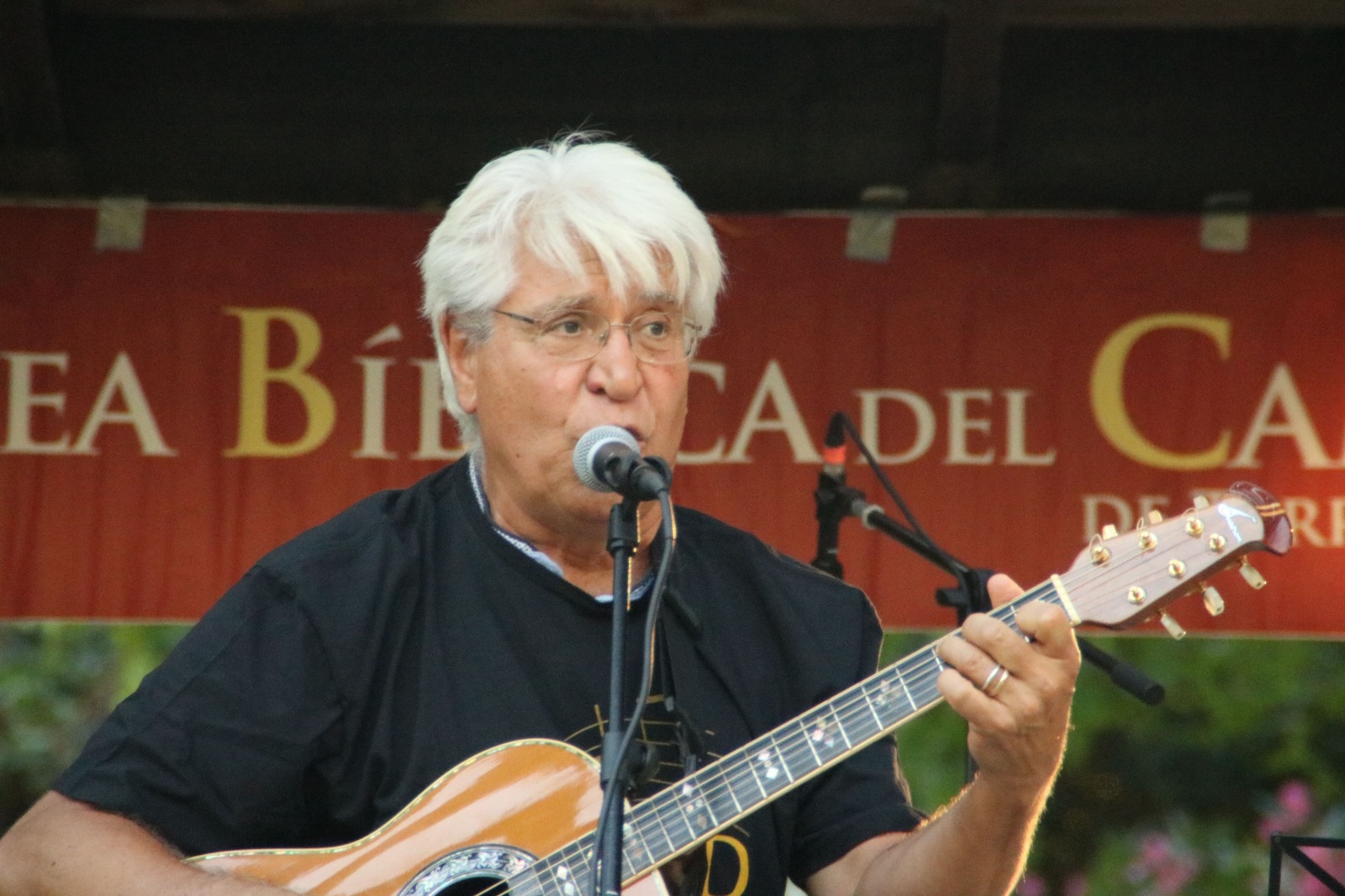 Primer plano de Joan Marc, con gafas y camiseta negra, cantando al micrófono y tocando la guitarra acústica ante el cartel rojo del escenario.