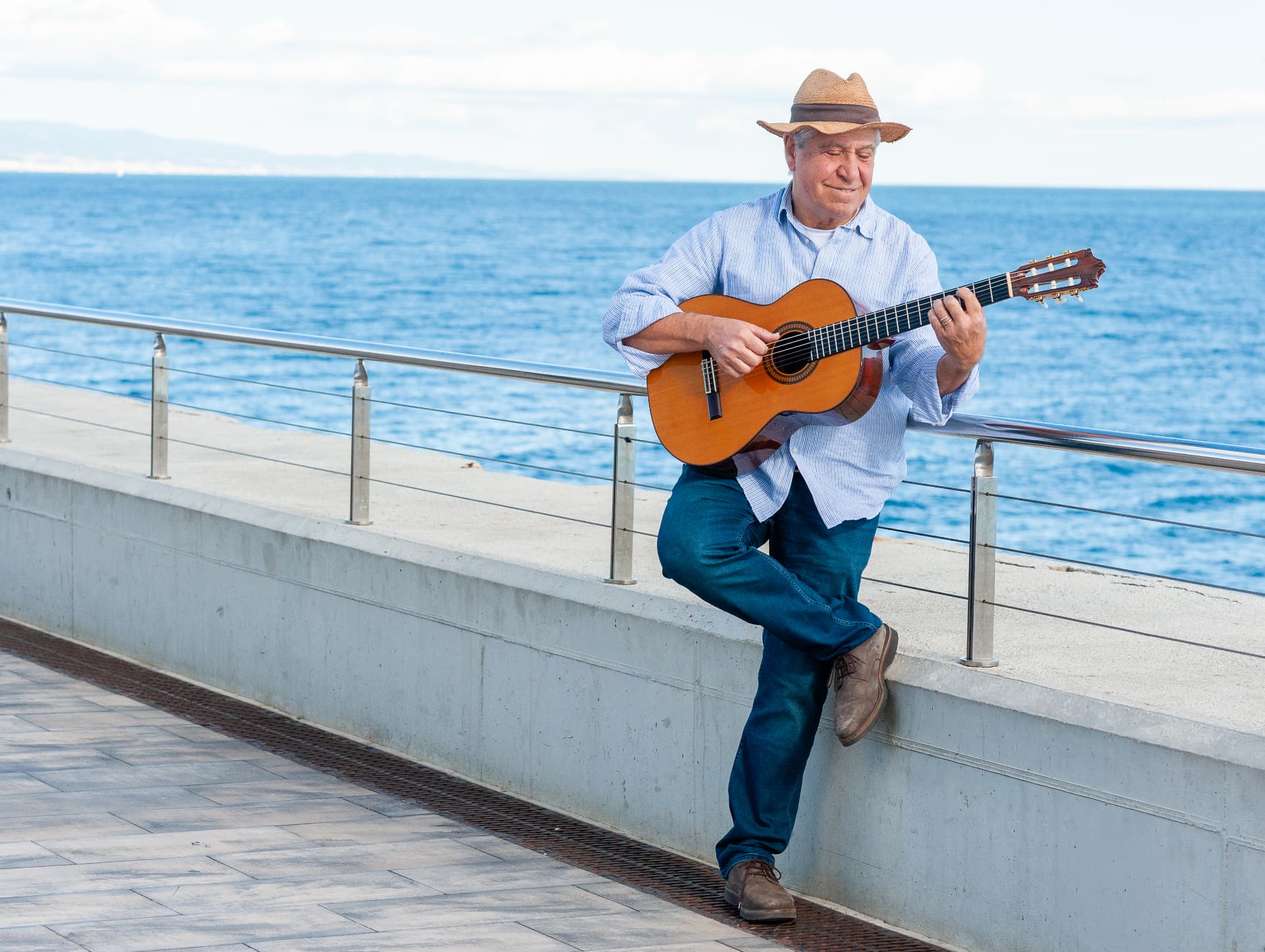 Con sombrero de paja y camisa azul clara, Joan Marc toca la guitarra apoyado en un muro del paseo marítimo, con las piernas cruzadas y el mar detrás.