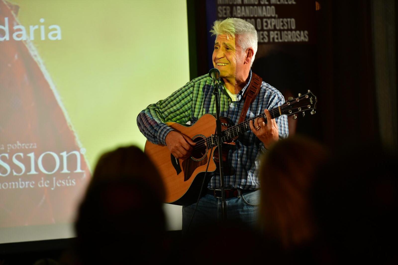 Joan Marc de pie, cantando y tocando la guitarra ante el micrófono, con el público en primer plano y una pantalla proyectada al fondo.