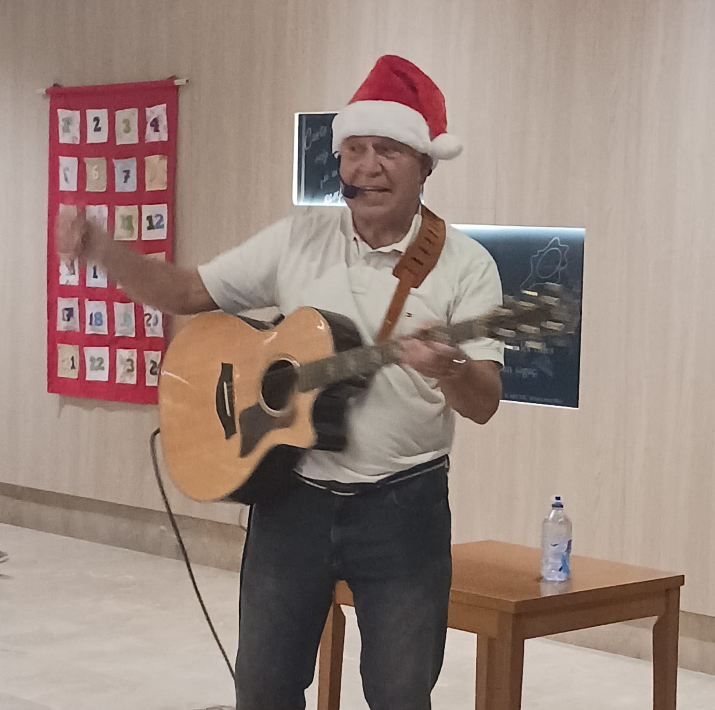 Joan Marc, con polo blanco y gorro de Papá Noel, canta con la guitarra y levanta una mano junto a un calendario de Adviento en el hospital.