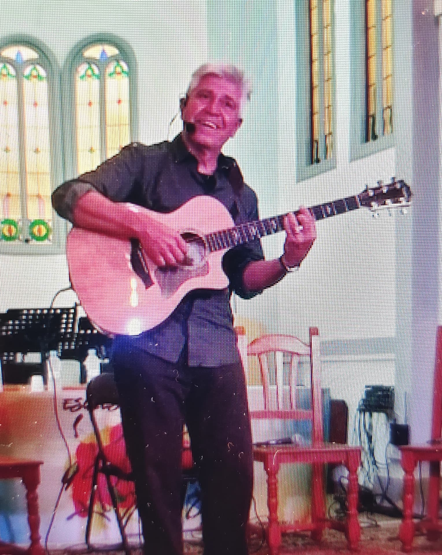 Joan Marc cantando con micrófono de diadema y guitarra acústica en una iglesia, con sillas rojas y un teclado al fondo.