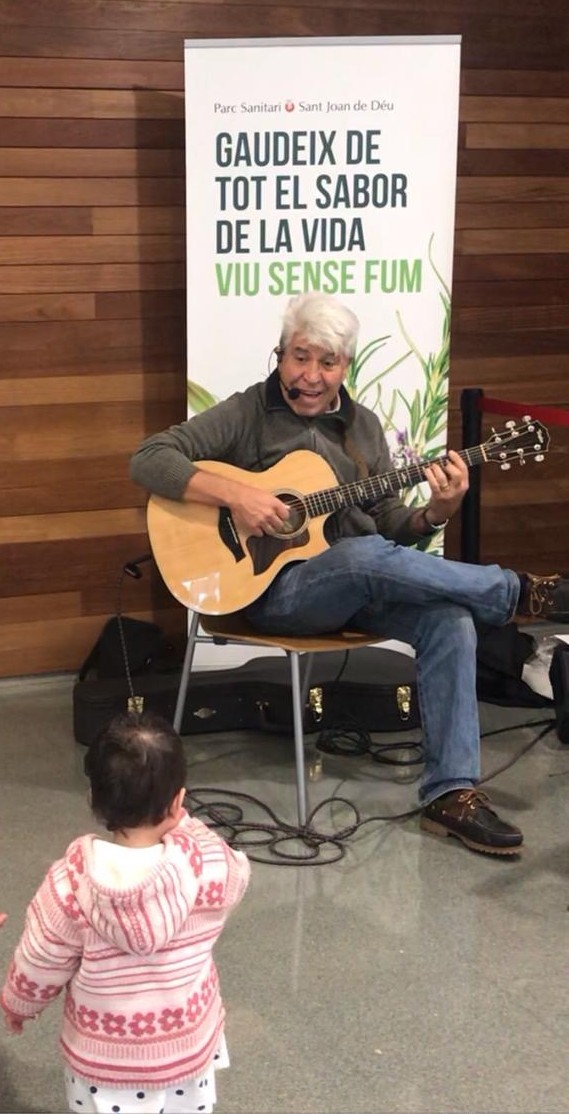 Joan Marc tocando la guitarra sentado en el pasillo del hospital, junto a la señal de Sortida y con personal sanitario de fondo.