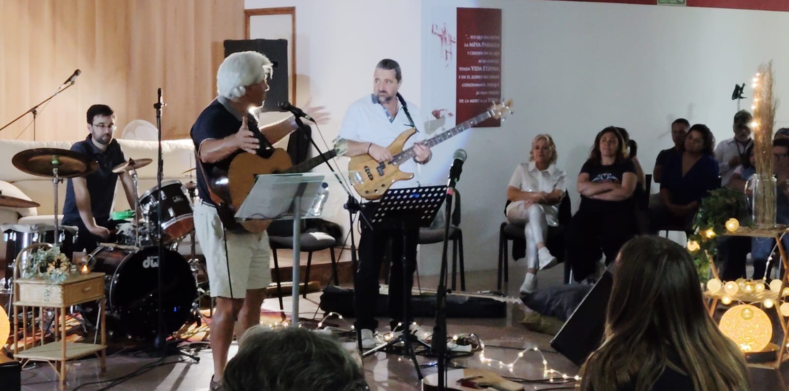 Joan Marc, con polo azul y guitarra acústica, canta junto a un bajista y un baterista en una sala de madera iluminada con luces cálidas; el público escucha sentado.