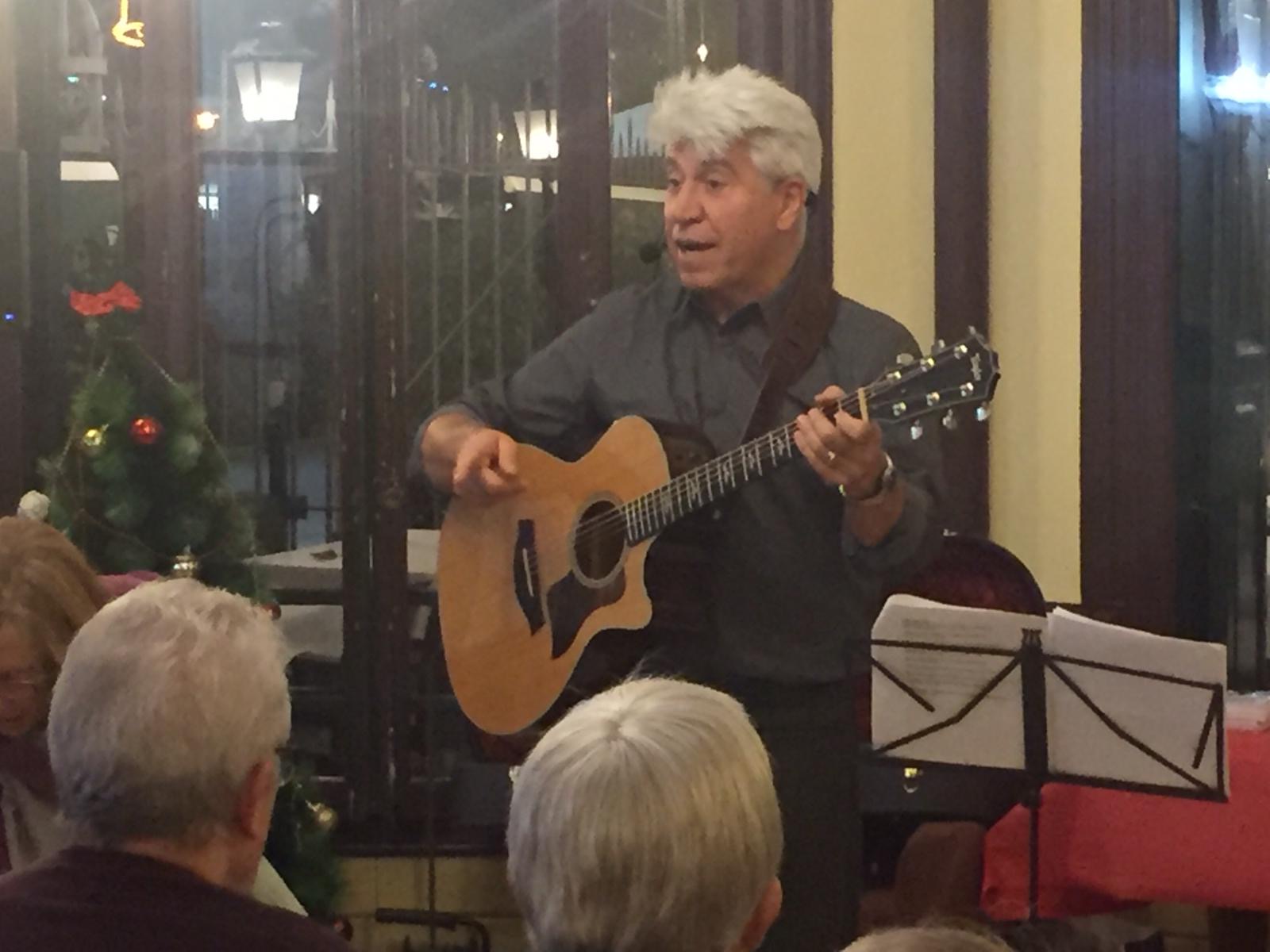 Joan Marc cantando con micrófono de diadema y guitarra en un interior con un pequeño árbol de Navidad y un atril con partituras.