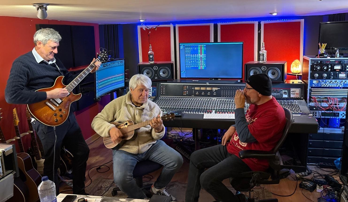Tres músicos comparten una sesión en el estudio: Joan Marc toca el ukelele sentado entre un guitarrista de pie y un músico con una armónica, ante la mesa de mezclas.