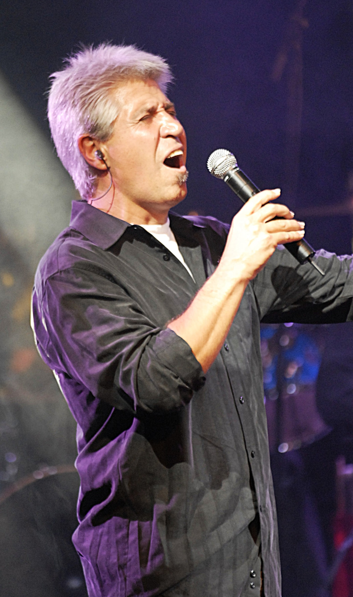 Primer plano de Joan Marc cantando con el micrófono, con el fondo oscuro salpicado de luces.