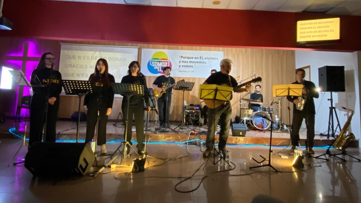 Sobre un escenario de madera, tres voces femeninas y una banda acompañan a Joan Marc, que canta con la guitarra en primer plano junto a un saxofonista.