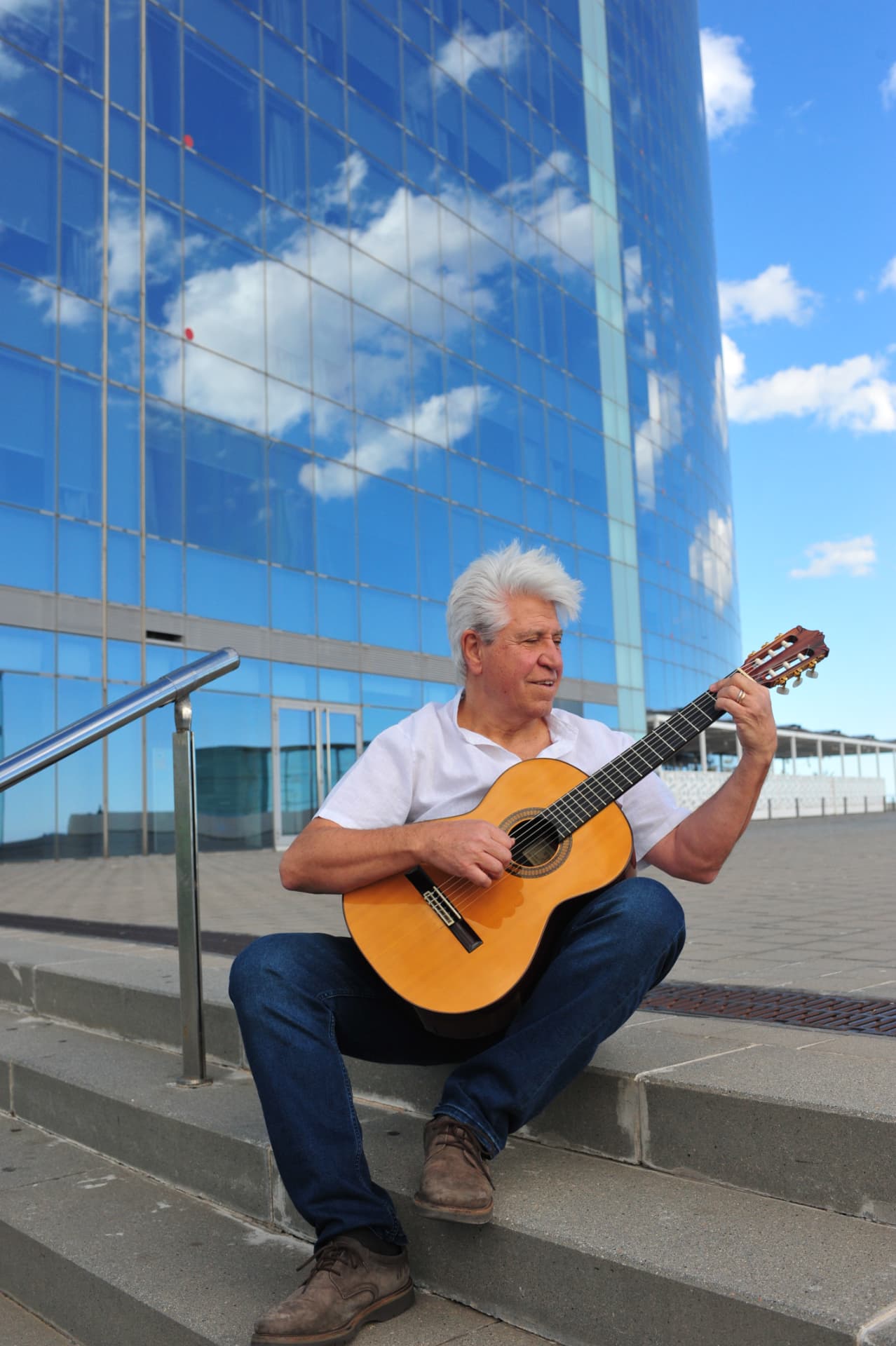 Joan Marc, con camisa blanca, toca la guitarra sentado en unas escaleras, ante un edificio acristalado que refleja el cielo y las nubes.