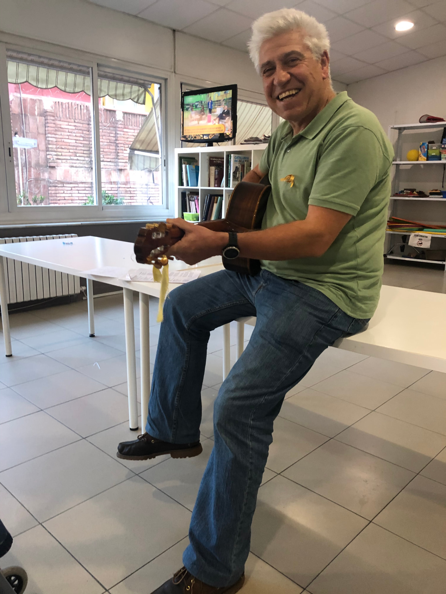 Joan Marc, sentado en el borde de una mesa con la guitarra y un lazo amarillo prendido al pecho, en una sala con ventanales y estanterías.