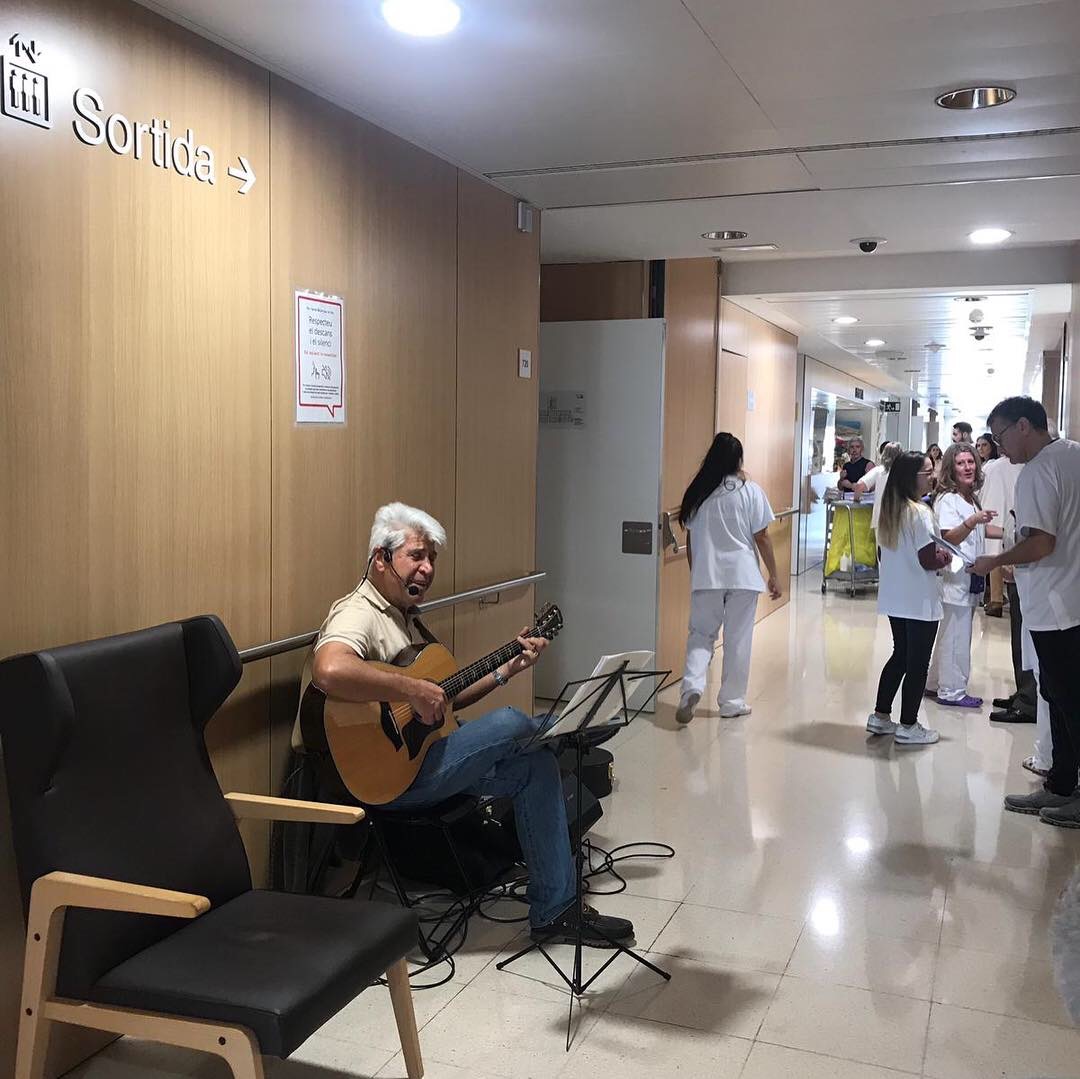 Joan Marc con la guitarra y el atril en el vestíbulo del hospital, junto al banner «Gaudeix de tot el sabor de la vida».