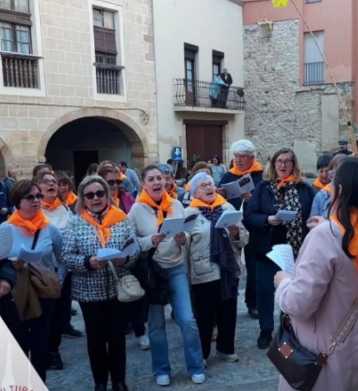 La Coral La Vaireta canta con partituras y pañuelos naranjas en una plaza de Alcover; Joan Marc aparece entre las voces, al fondo.