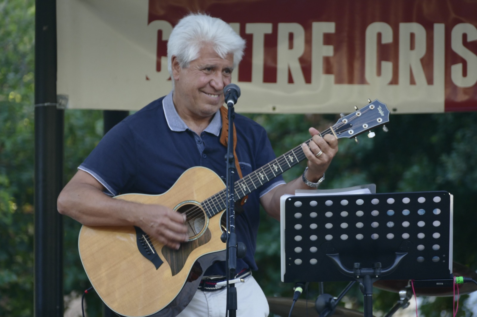 Joan Marc, con polo azul marino, sonríe al micrófono mientras toca la guitarra acústica en un escenario al aire libre, con un atril negro delante.