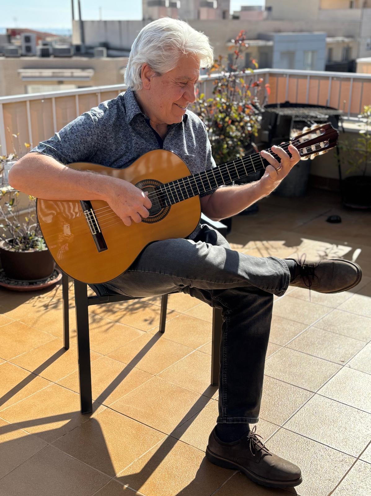 Joan Marc sonríe mientras toca la guitarra clásica sentado con las piernas cruzadas en una terraza soleada, entre macetas y edificios.