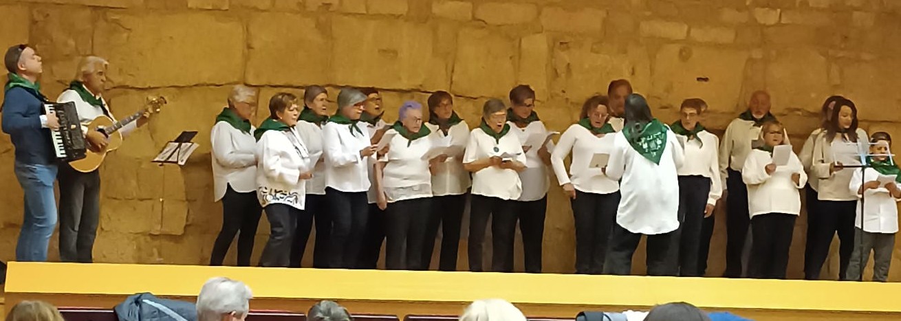 Panorámica del coro cantando ante un muro de piedra antigua, dirigido por una maestra vista de espaldas, con un acordeón y una guitarra a la izquierda.