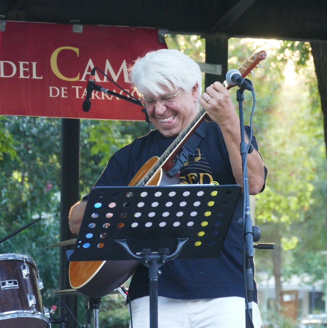 Plano cercano de Joan Marc sonriendo mientras toca la guitarra al aire libre, con un atril perforado delante y el cartel rojo del escenario detrás.