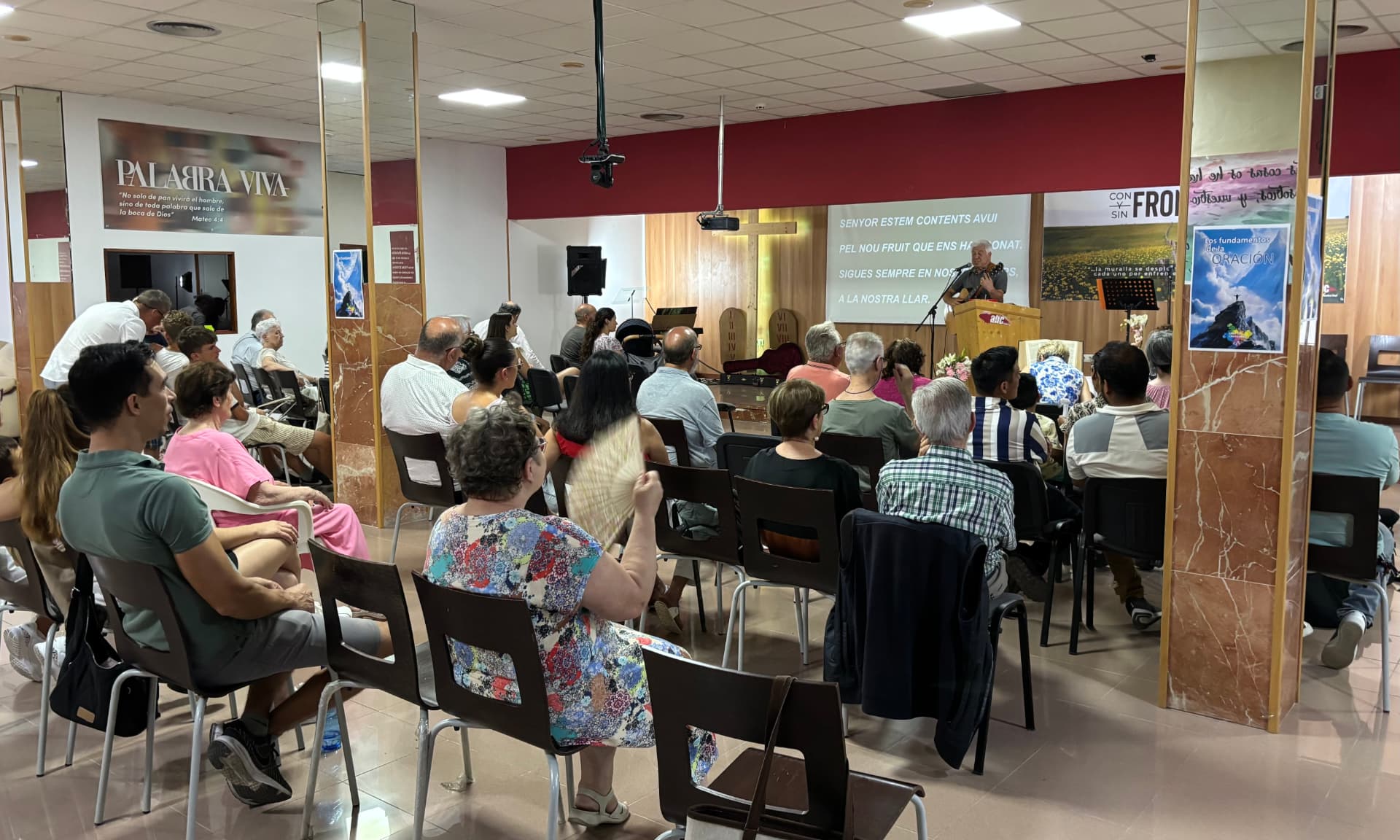 La asamblea sentada en filas mira hacia el escenario, donde Joan Marc canta y toca la guitarra detrás de un atril de madera.