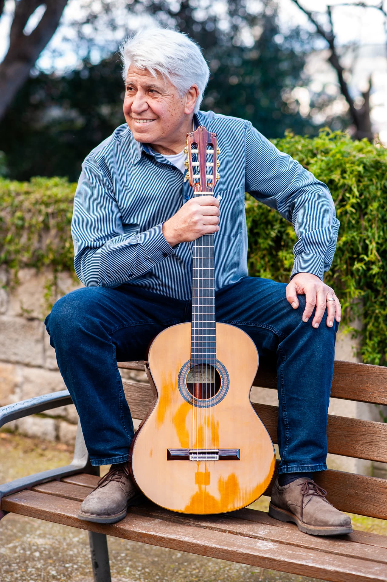 Joan Marc sonríe sentado en un banco de madera, con la guitarra clásica apoyada verticalmente entre las piernas y un jardín detrás.