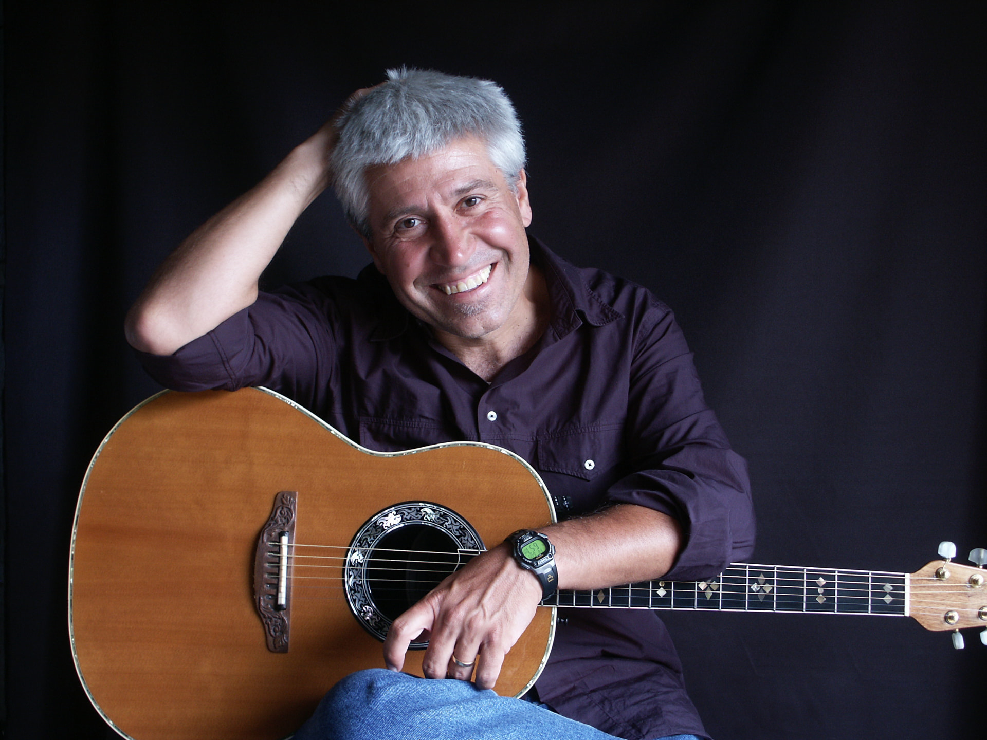 Joan Marc sonriendo, con una mano sobre la cabeza y una guitarra acústica apoyada sobre las piernas.