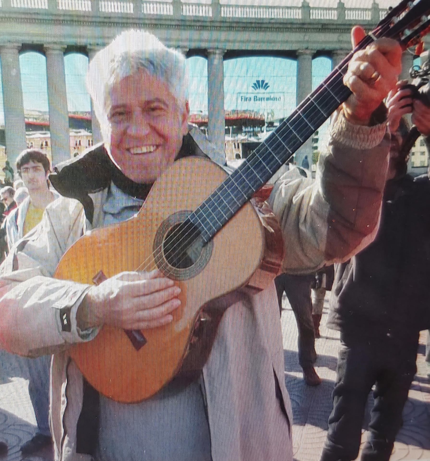 Joan Marc de pie en una plaza, sosteniendo en alto una guitarra clásica, con transeúntes y una columnata al fondo.