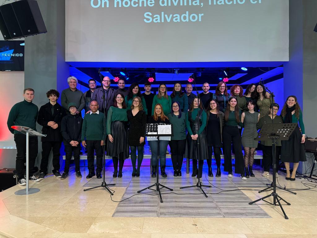 Veinticinco voces del coro posan en el escenario en filas escalonadas, con jerseys verdes y ropa oscura, bajo una luz azul.
