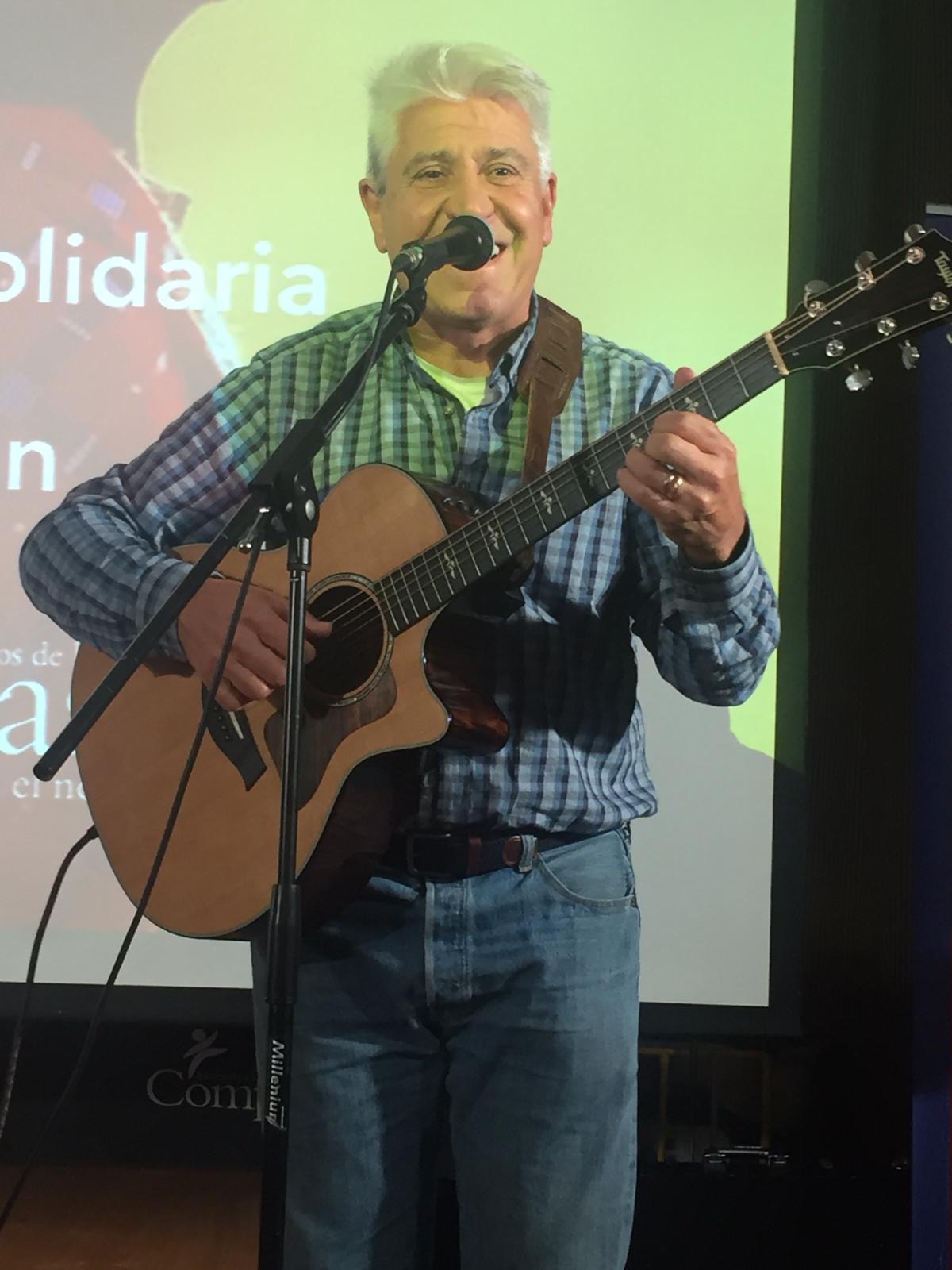 Joan Marc, sonriente y con los ojos entrecerrados, canta y toca la guitarra frente al micrófono, ante una pantalla de proyección.