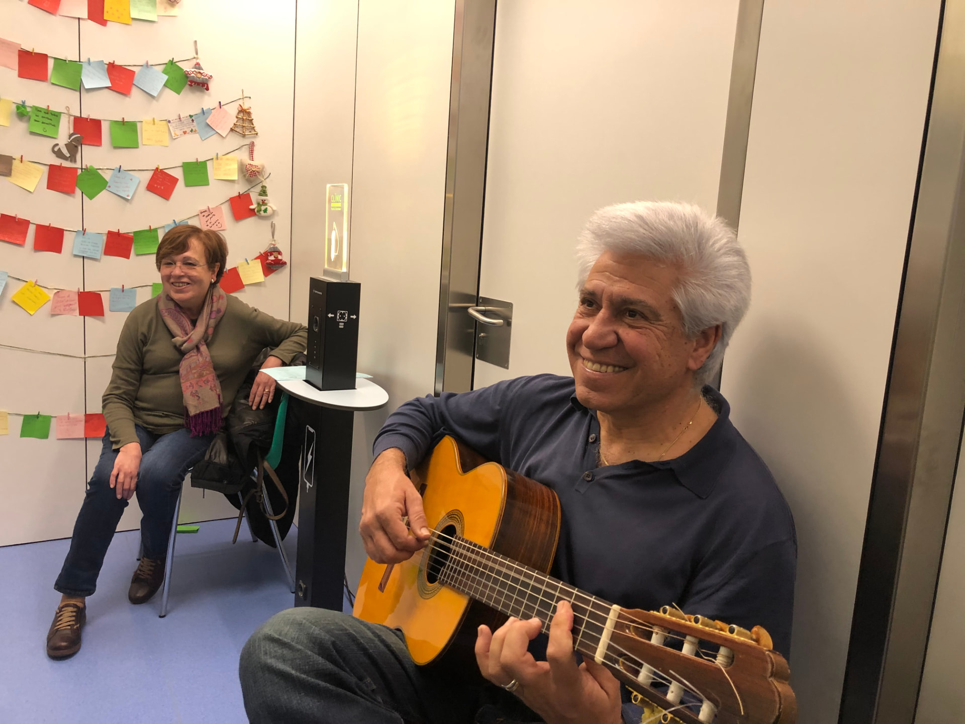 Joan Marc sonriendo con la guitarra clásica en una sala del hospital, junto a una mujer sentada y un árbol de notas de colores.
